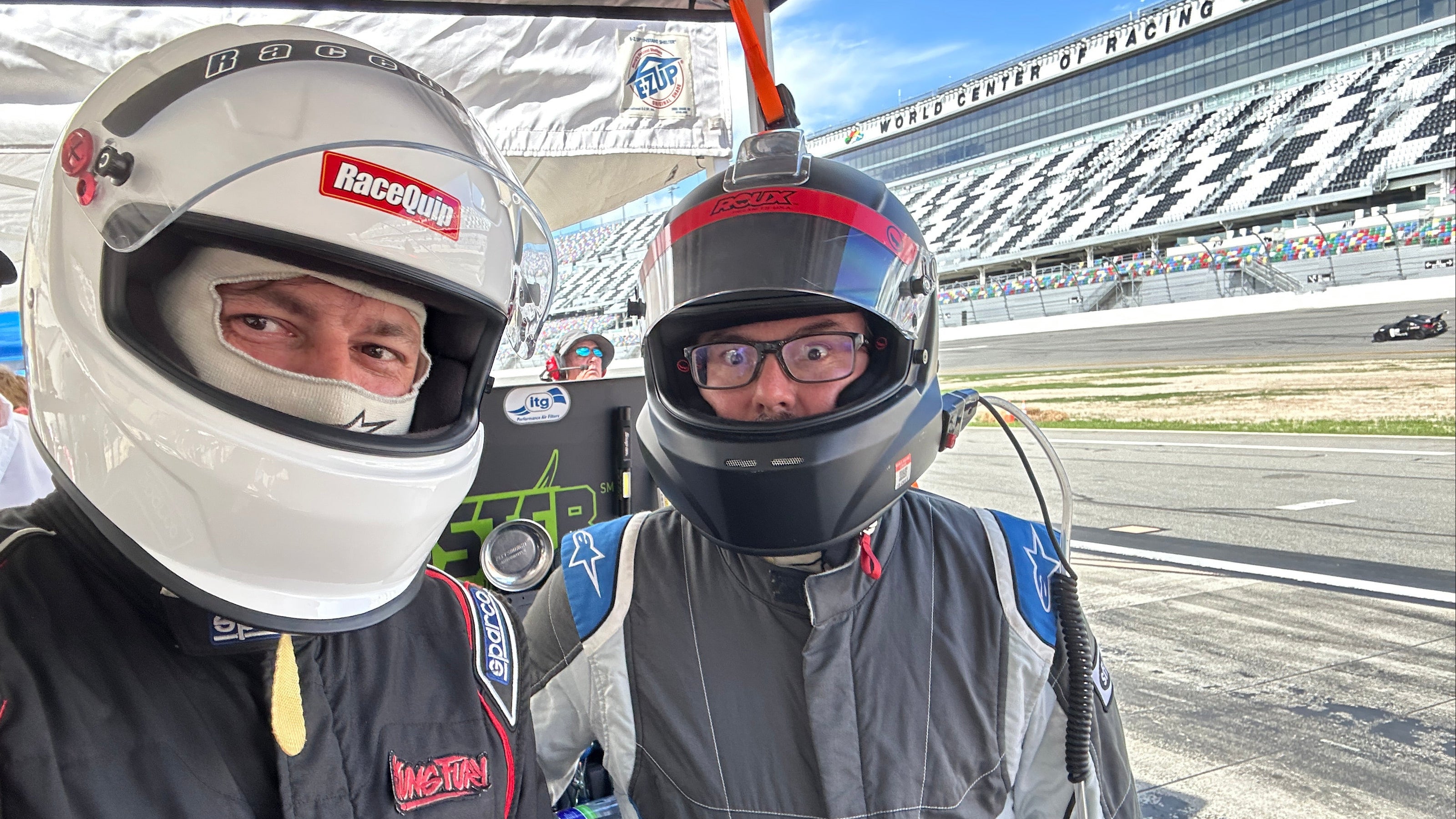 Two race drivers in full racing gear and helmets standing under a canopy at a racetrack. One driver wears a white helmet and black suit with red and white sponsor logos, and the other wears a black helmet and gray suit with blue accents.