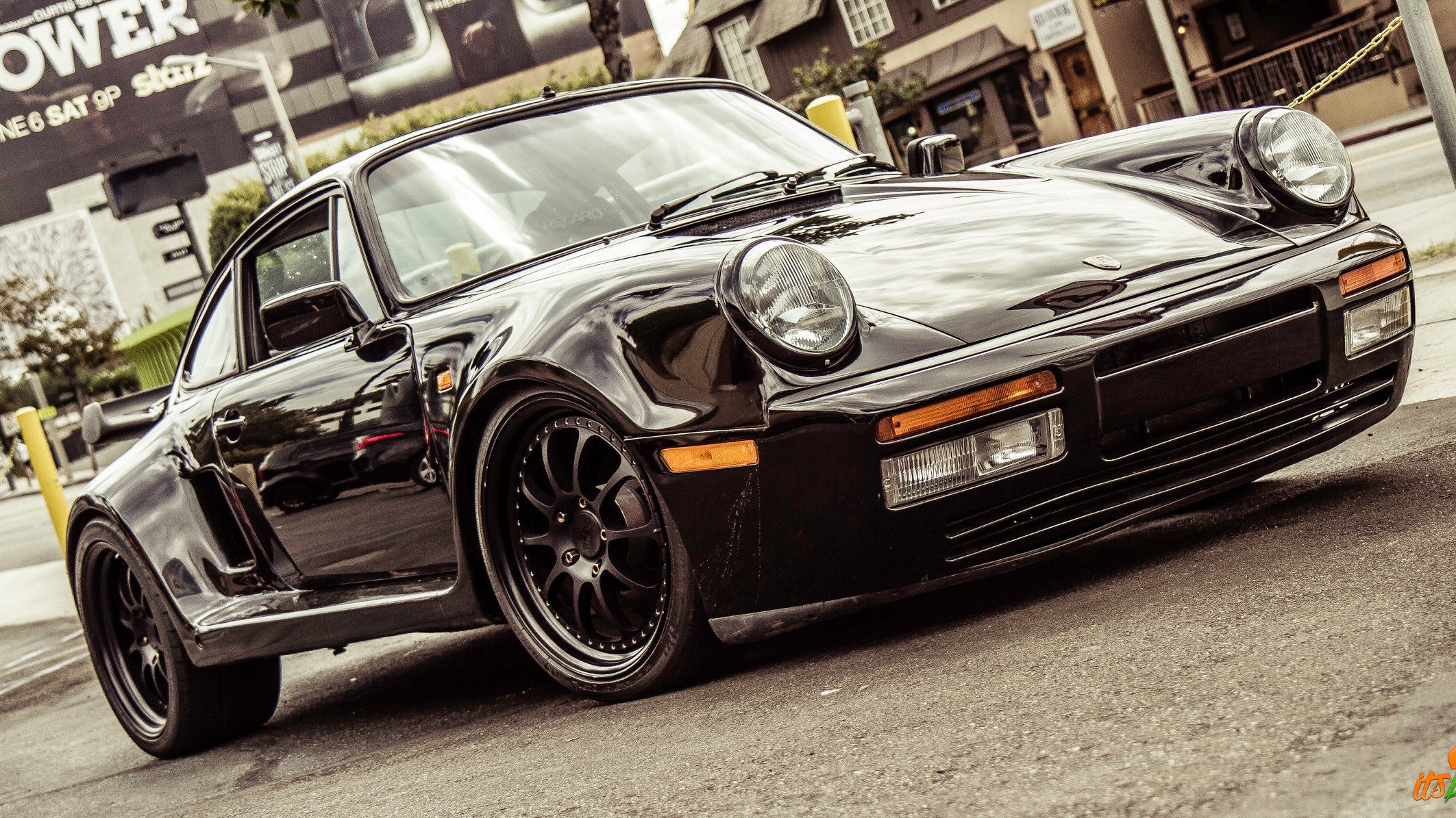 A black Porsche 911 with widebody styling and black wheels, photographed on a city street with buildings and a billboard in the background.