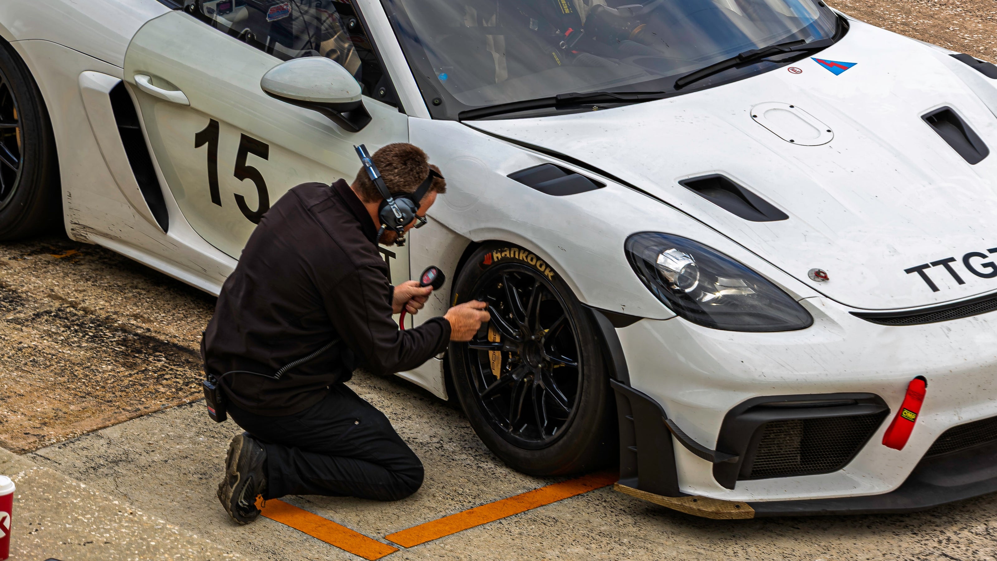 A white Porsche 911 GT3 race car on a racetrack with a mechanic kneeling beside the front wheel, working on the tire. The car has the number 15 on the door and 'TTG' on the hood.